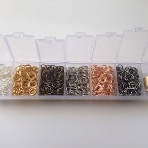 May include: A clear plastic storage box with six compartments filled with silver, gold, bronze, and black jump rings. A gold metal ring is in the last compartment.