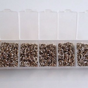 May include: A clear plastic storage box with eight compartments filled with silver jump rings.
