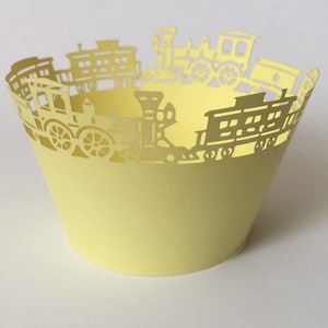 May include: Yellow cupcake liners with a cut-out design of a train engine and boxcars. The design is intricate and detailed, with wheels, windows, and smoke stacks.