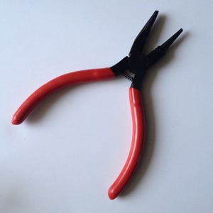 May include: Round-nose pliers with red handles and black jaws. The pliers have a spring and are designed for precision work. The tool is set against a white background.