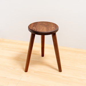 May include: A round wooden stool with three legs. The stool is made of walnut wood and has a natural finish. The top of the stool has a brass inlay.