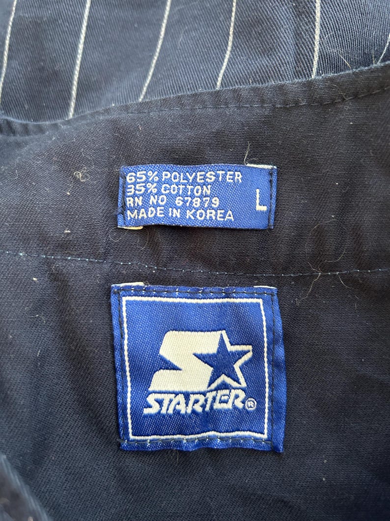 Pu&ograve; includere: Primo piano di una camicia blu navy con sottili righe bianche. La camicia ha due etichette: una con il logo Starter e un'altra con la scritta "65% Polyester 35% Cotton RN NO 67879 MADE IN KOREA" e la taglia "L".