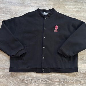 May include: Black varsity jacket with a button-up front and a stand-up collar. The jacket features a red and white embroidered logo on the chest. The jacket has two front pockets and ribbed cuffs and hem. The jacket is made of a wool blend.
