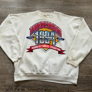 May include: White sweatshirt with a graphic that reads "Thunderbird 1994 Manufacturers Champion." The graphic is in red, blue, and yellow. The sweatshirt has long sleeves and a crew neck.