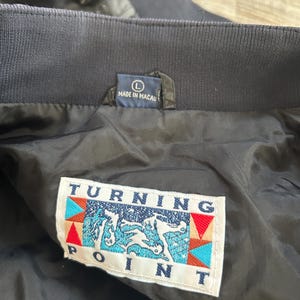 Pode incluir: Close-up do interior de um casaco azul marinho. O casaco tem uma gola canelada e um forro preto. Uma etiqueta diz "MADE IN MACAU" e outra etiqueta exibe o texto "TURNING POINT" com um gr&aacute;fico de um cavalo.