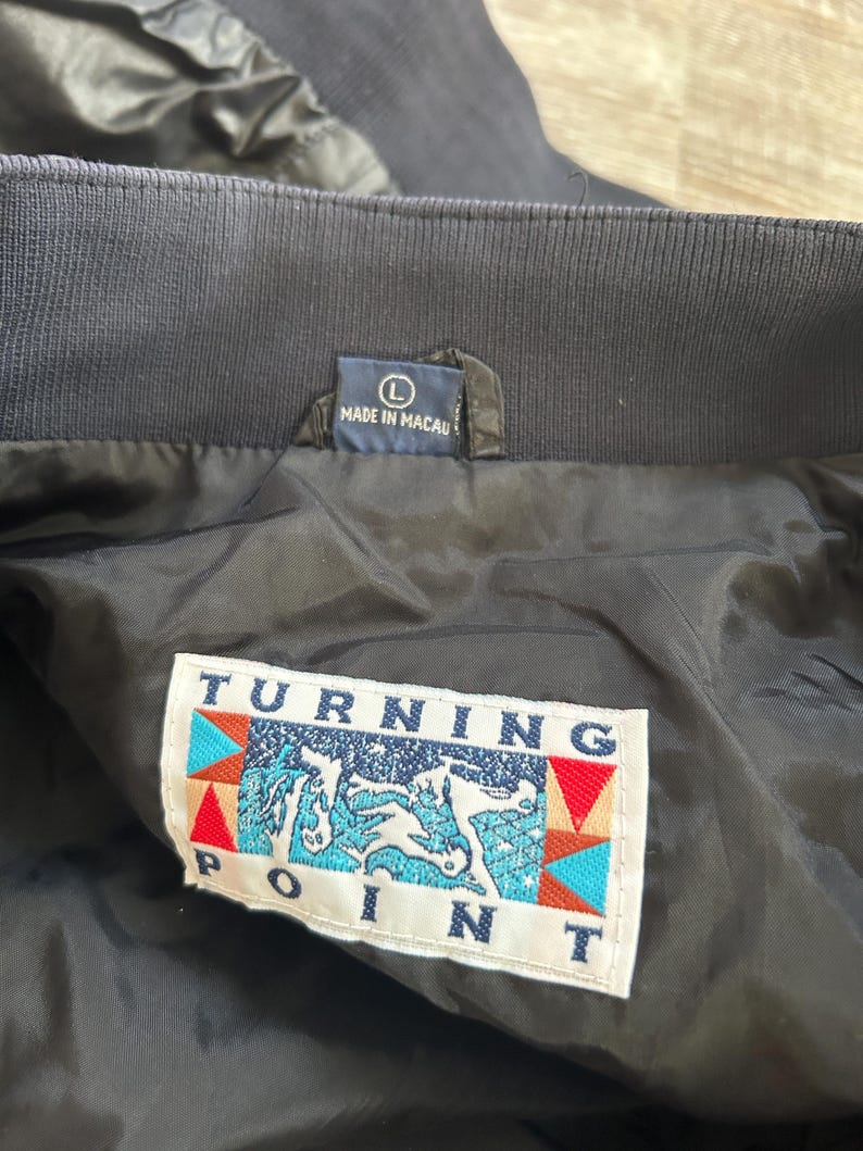 Pode incluir: Close-up do interior de um casaco azul marinho. O casaco tem uma gola canelada e um forro preto. Uma etiqueta diz "MADE IN MACAU" e outra etiqueta exibe o texto "TURNING POINT" com um gr&aacute;fico de um cavalo.