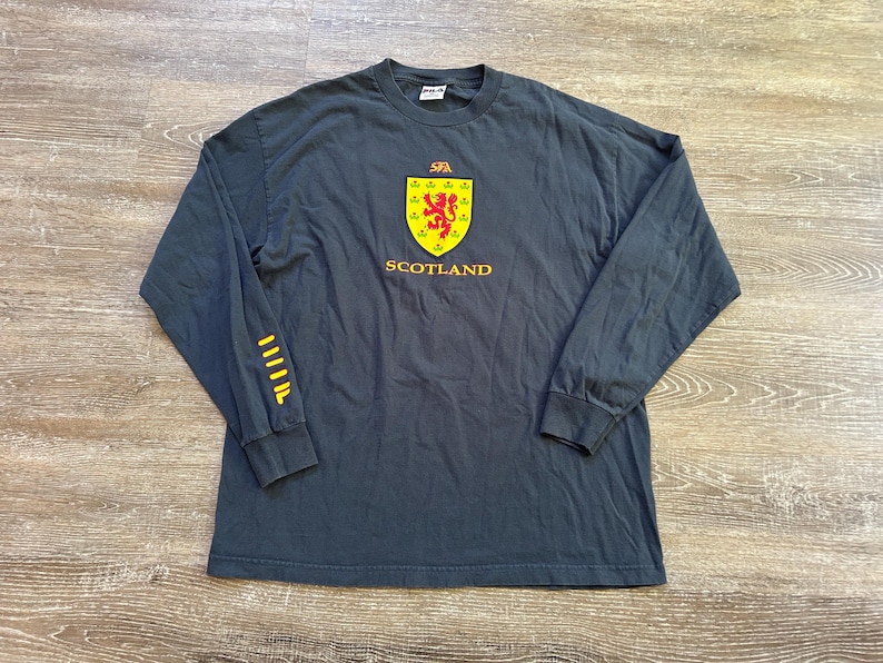 Peut inclure: T-shirt &agrave; manches longues gris fonc&eacute; avec l'embl&egrave;me de l'&Eacute;cosse. L'embl&egrave;me est un &eacute;cusson jaune avec un lion rouge et le mot "SCOTLAND" en jaune. La manche gauche a des rayures jaunes. Le t-shirt est en mati&egrave;re souple.
