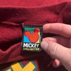 May include: Close-up of a burgundy sweatshirt with a Mickey Unlimited tag. The tag features a red Mickey Mouse head on a blue background, with the text "MICKEY UNLIMITED" in white.