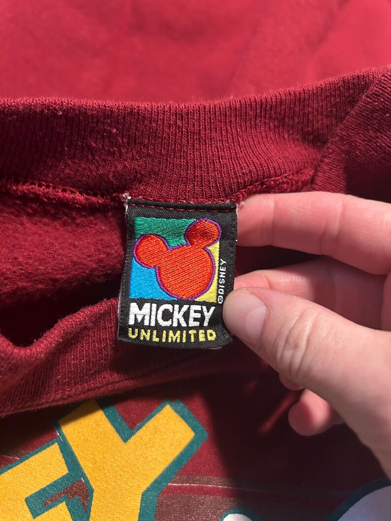 May include: Close-up of a burgundy sweatshirt with a Mickey Unlimited tag. The tag features a red Mickey Mouse head on a blue background, with the text "MICKEY UNLIMITED" in white.