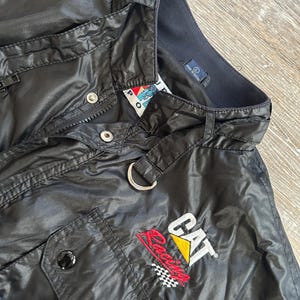 Pode incluir: Casaco preto de nylon com fecho de correr, bot&otilde;es de press&atilde;o e um pequeno bolso. O casaco apresenta o log&oacute;tipo "CAT Racing" em vermelho, amarelo e branco, com um design de bandeira quadriculada. Um anel de metal prateado est&aacute; preso &agrave; gola.