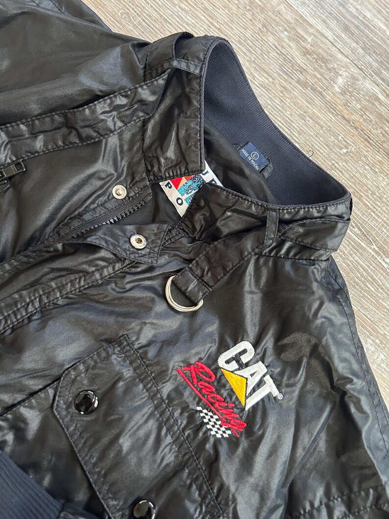 Pode incluir: Casaco preto de nylon com fecho de correr, bot&otilde;es de press&atilde;o e um pequeno bolso. O casaco apresenta o log&oacute;tipo "CAT Racing" em vermelho, amarelo e branco, com um design de bandeira quadriculada. Um anel de metal prateado est&aacute; preso &agrave; gola.