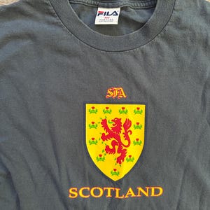 Peut inclure: T-shirt FILA gris fonc&eacute; avec un blason &eacute;cossais jaune et rouge. Le blason pr&eacute;sente un lion rouge rampant et le mot "SCOTLAND" en orange. Le t-shirt a un col rond et une &eacute;tiquette FILA.