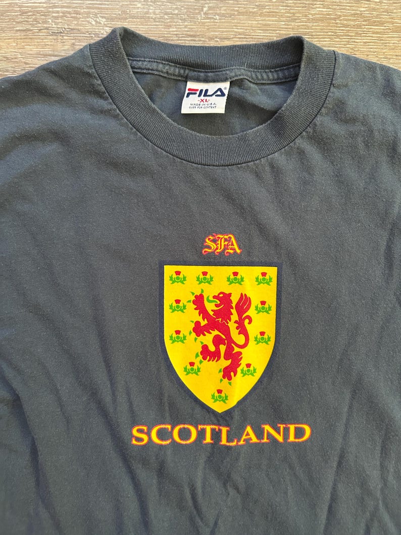 Peut inclure: T-shirt FILA gris fonc&eacute; avec un blason &eacute;cossais jaune et rouge. Le blason pr&eacute;sente un lion rouge rampant et le mot "SCOTLAND" en orange. Le t-shirt a un col rond et une &eacute;tiquette FILA.