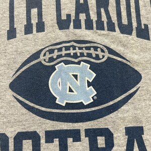 Może przedstawiać: Szara koszulka z napisem "NORTH CAROLINA FOOTBALL" w granatowych literach. Granatowa grafika piłki nożnej z jasnoniebieskim logo "NC" jest wyśrodkowana na koszulce.