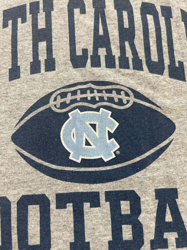 Może przedstawiać: Szara koszulka z napisem "NORTH CAROLINA FOOTBALL" w granatowych literach. Granatowa grafika piłki nożnej z jasnoniebieskim logo "NC" jest wyśrodkowana na koszulce.