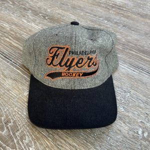 May include: A gray and black baseball cap featuring the Philadelphia Flyers hockey team logo in orange and black embroidery. The cap has a curved brim and a small black button on top.