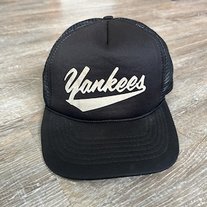May include: Black trucker hat with the word "Yankees" in white script across the front. The hat has a mesh back and a curved brim. The hat is sitting on a wooden surface.