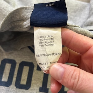 Może przedstawiać: Zbliżenie szarej koszulki z granatową metką. Na metce widnieje napis "90% Bawełna, 10% Poliester" oraz "Adult 2XL". Widoczny jest materiał koszulki, a słowo "FOOT" jest częściowo widoczne w kolorze granatowym.