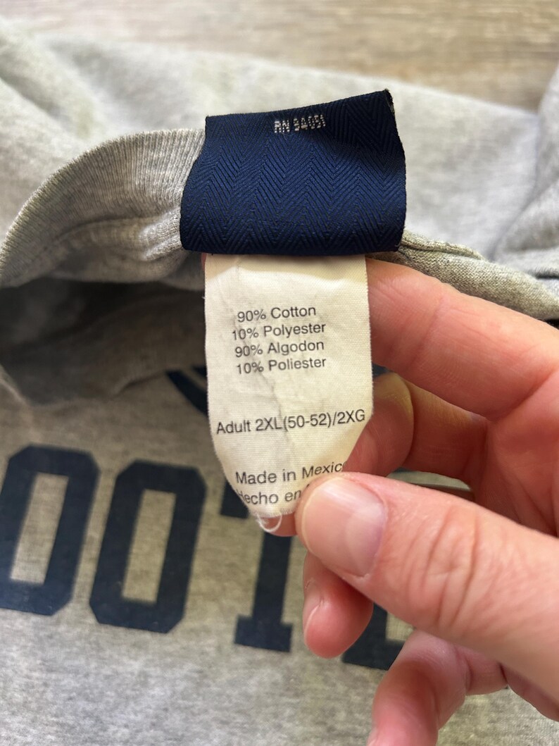Może przedstawiać: Zbliżenie szarej koszulki z granatową metką. Na metce widnieje napis "90% Bawełna, 10% Poliester" oraz "Adult 2XL". Widoczny jest materiał koszulki, a słowo "FOOT" jest częściowo widoczne w kolorze granatowym.