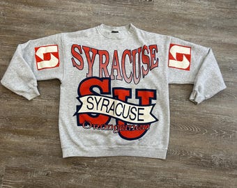 May include: A gray sweatshirt with long sleeves featuring the word "SYRACUSE" in red and white lettering. The sweatshirt also has the word "SYRACUSE" on a banner and the word "Orangemen" in red. The sleeves have a red and white logo.