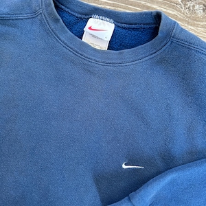 May include: A blue sweatshirt with a white Nike swoosh logo on the front. The sweatshirt has a crew neck and long sleeves. The tag on the sweatshirt says "Nike" and "Made in USA".