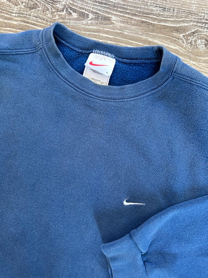May include: A blue sweatshirt with a white Nike swoosh logo on the front. The sweatshirt has a crew neck and long sleeves. The tag on the sweatshirt says "Nike" and "Made in USA".