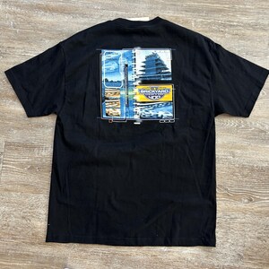 May include: A black short-sleeve t-shirt featuring a graphic on the back. The design includes a collage of images showing a race track, a tall structure, and a yellow rectangular logo with the text Brickyard 400 printed in blue and white.