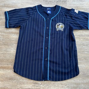 Pu&ograve; includere: Maglia da baseball blu navy con gessati bianchi e finiture azzurre. Presenta una parte anteriore con bottoni, maniche corte e una toppa con il logo della squadra sul petto. Il logo Starter &egrave; visibile sulla manica.