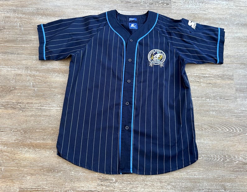 Pu&ograve; includere: Maglia da baseball blu navy con gessati bianchi e finiture azzurre. Presenta una parte anteriore con bottoni, maniche corte e una toppa con il logo della squadra sul petto. Il logo Starter &egrave; visibile sulla manica.