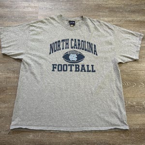 Może przedstawiać: Szara koszulka z napisem "NORTH CAROLINA FOOTBALL" w kolorze granatowym. Słowo "NORTH CAROLINA" jest wygięte nad grafiką piłki nożnej z logo UNC. Koszulka jest wykonana z miękkiego materiału.