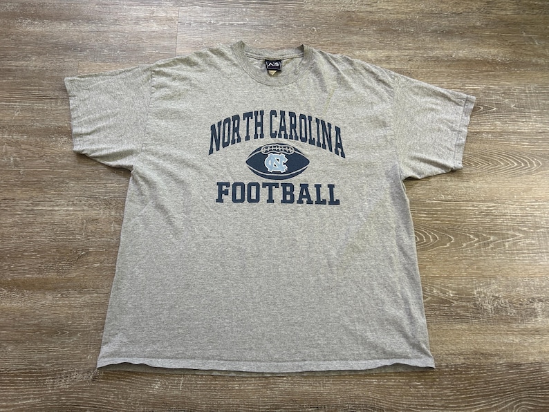 Może przedstawiać: Szara koszulka z napisem "NORTH CAROLINA FOOTBALL" w kolorze granatowym. Słowo "NORTH CAROLINA" jest wygięte nad grafiką piłki nożnej z logo UNC. Koszulka jest wykonana z miękkiego materiału.