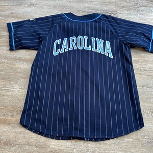 Pu&ograve; includere: Maglia da baseball blu navy con gessati bianchi e la scritta "CAROLINA" in azzurro sul petto. La maglia ha maniche corte con finiture azzurre e un logo Starter sulla manica sinistra.