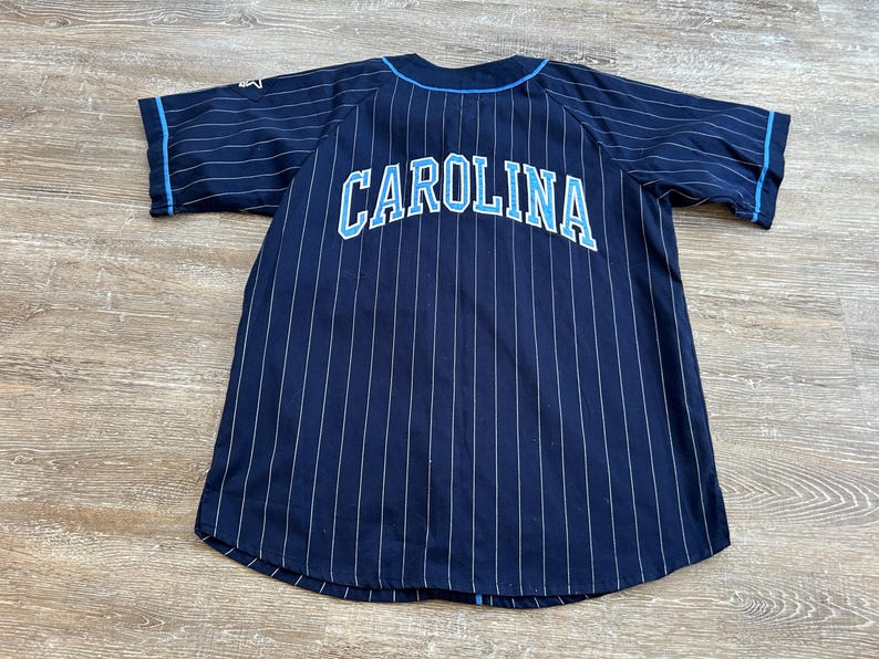 Pu&ograve; includere: Maglia da baseball blu navy con gessati bianchi e la scritta "CAROLINA" in azzurro sul petto. La maglia ha maniche corte con finiture azzurre e un logo Starter sulla manica sinistra.