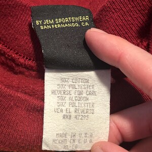 May include: Close-up of a burgundy sweatshirt tag. The tag reads "BY JEM SPORTSWEAR, SAN FERNANDO, CA." and lists fabric content as 50% cotton and 50% polyester. The tag also includes care instructions and "MADE IN U.S.A."