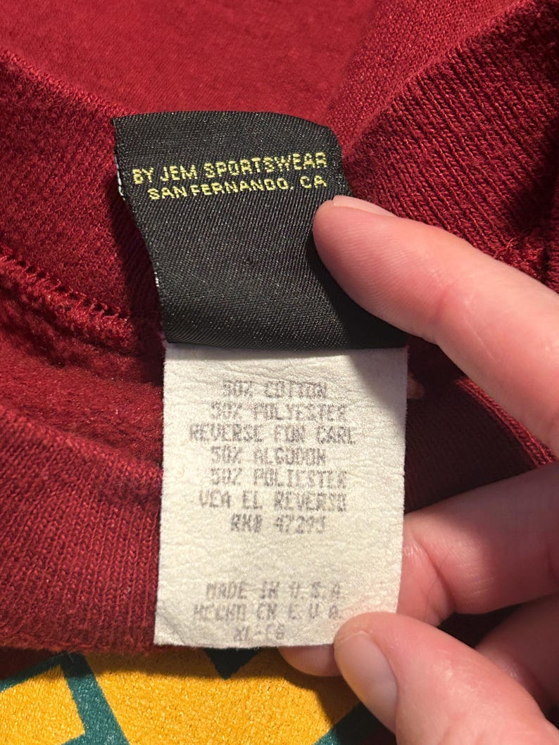 May include: Close-up of a burgundy sweatshirt tag. The tag reads "BY JEM SPORTSWEAR, SAN FERNANDO, CA." and lists fabric content as 50% cotton and 50% polyester. The tag also includes care instructions and "MADE IN U.S.A."