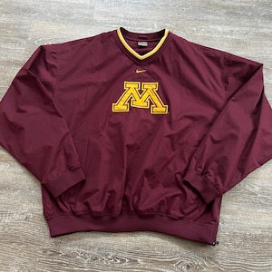 May include: Maroon pullover windbreaker with a yellow collar and the University of Minnesota "M" logo in yellow. The Nike logo is visible on the chest. The jacket has a V-neck and elastic cuffs.