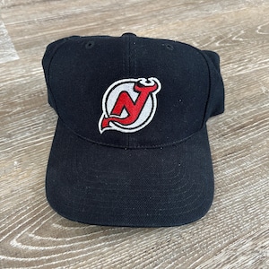 May include: Black baseball cap featuring the New Jersey Devils logo. The logo is a red "N" with a devil's tail, outlined in white, and set against a white circle. The cap is made of a durable fabric and has a curved brim.