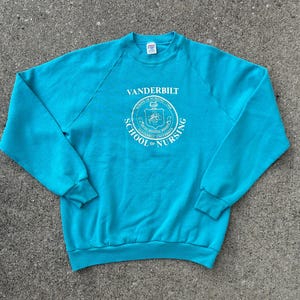 May include: A bright turquoise long-sleeve sweatshirt featuring a white circular graphic on the chest. The text reads VANDERBILT SCHOOL OF NURSING with a central emblem. The garment has a crew neckline, raglan sleeves, and ribbed cuffs and hem.