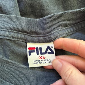 Peut inclure: Gros plan sur un col de t-shirt gris fonc&eacute; avec une &eacute;tiquette blanche. L'&eacute;tiquette pr&eacute;sente le logo FILA en rouge et bleu marine, avec le texte "-XL- MADE IN U.S.A. OVER FOR CONTENT".