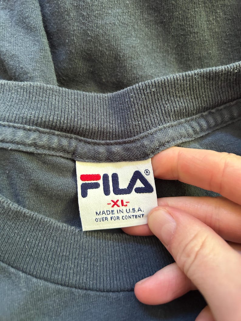 Peut inclure: Gros plan sur un col de t-shirt gris fonc&eacute; avec une &eacute;tiquette blanche. L'&eacute;tiquette pr&eacute;sente le logo FILA en rouge et bleu marine, avec le texte "-XL- MADE IN U.S.A. OVER FOR CONTENT".