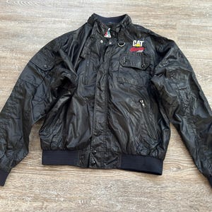 Pode incluir: Casaco de nylon preto com gola alta, dois bolsos no peito e o log&oacute;tipo CAT no peito direito. O casaco tem fecho de correr e punhos e bainha canelados. O casaco &eacute; tamanho m&eacute;dio.