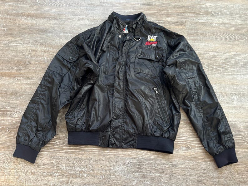 Pode incluir: Casaco de nylon preto com gola alta, dois bolsos no peito e o log&oacute;tipo CAT no peito direito. O casaco tem fecho de correr e punhos e bainha canelados. O casaco &eacute; tamanho m&eacute;dio.