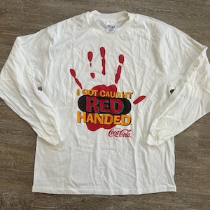 May include: White long-sleeve t-shirt with a red handprint graphic and the text "I got caught red handed" with the Coca-Cola logo.