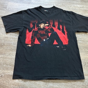 May include: Black t-shirt featuring a graphic of a person in a red and black plaid shirt and the name "Garth" in red block letters. The person is wearing a black hat and blue jeans. The shirt is on a light wood surface.