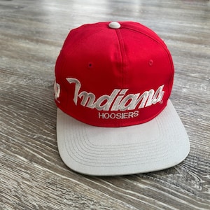 May include: A red and gray baseball cap with the word "Indians" embroidered in white script across the front. The word "HOOSIERS" is embroidered in white below the word "Indians". The cap has a white button on top.