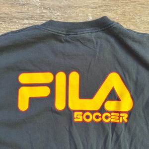 Peut inclure: Un t-shirt noir avec le logo FILA en jaune avec un contour rouge. Le mot "SOCCER" est imprim&eacute; en jaune avec un contour rouge en dessous du logo. Le t-shirt est fait d'une mati&egrave;re douce.
