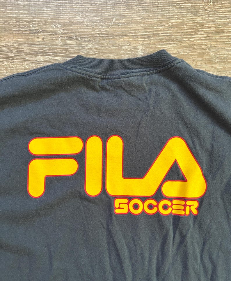Peut inclure: Un t-shirt noir avec le logo FILA en jaune avec un contour rouge. Le mot "SOCCER" est imprim&eacute; en jaune avec un contour rouge en dessous du logo. Le t-shirt est fait d'une mati&egrave;re douce.
