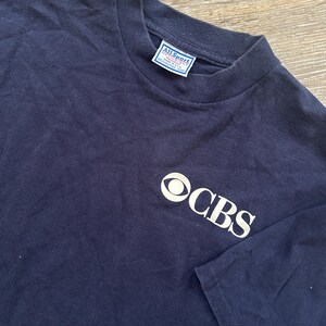 May include: Navy blue t-shirt with the CBS logo in white on the front. The shirt has a crew neck and short sleeves. The All Sport tag is visible on the inside of the collar.