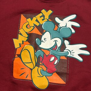May include: Maroon sweatshirt featuring a vintage Mickey Mouse graphic. The design includes Mickey in a dynamic pose with teal ears, red shorts, yellow shoes, and white gloves. The word "MICKEY" is in yellow block letters.