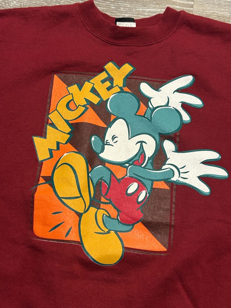 May include: Maroon sweatshirt featuring a vintage Mickey Mouse graphic. The design includes Mickey in a dynamic pose with teal ears, red shorts, yellow shoes, and white gloves. The word "MICKEY" is in yellow block letters.